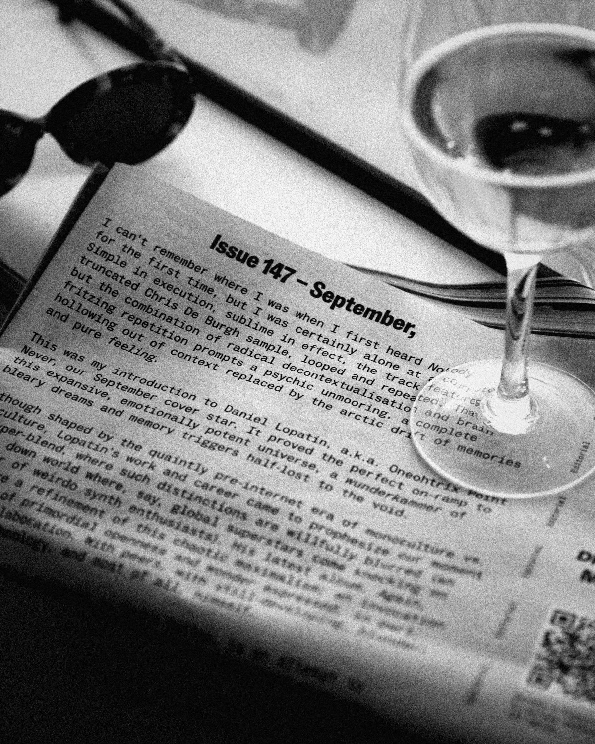 A printed magazine page next to sunglasses and a glass of white wine, black and white