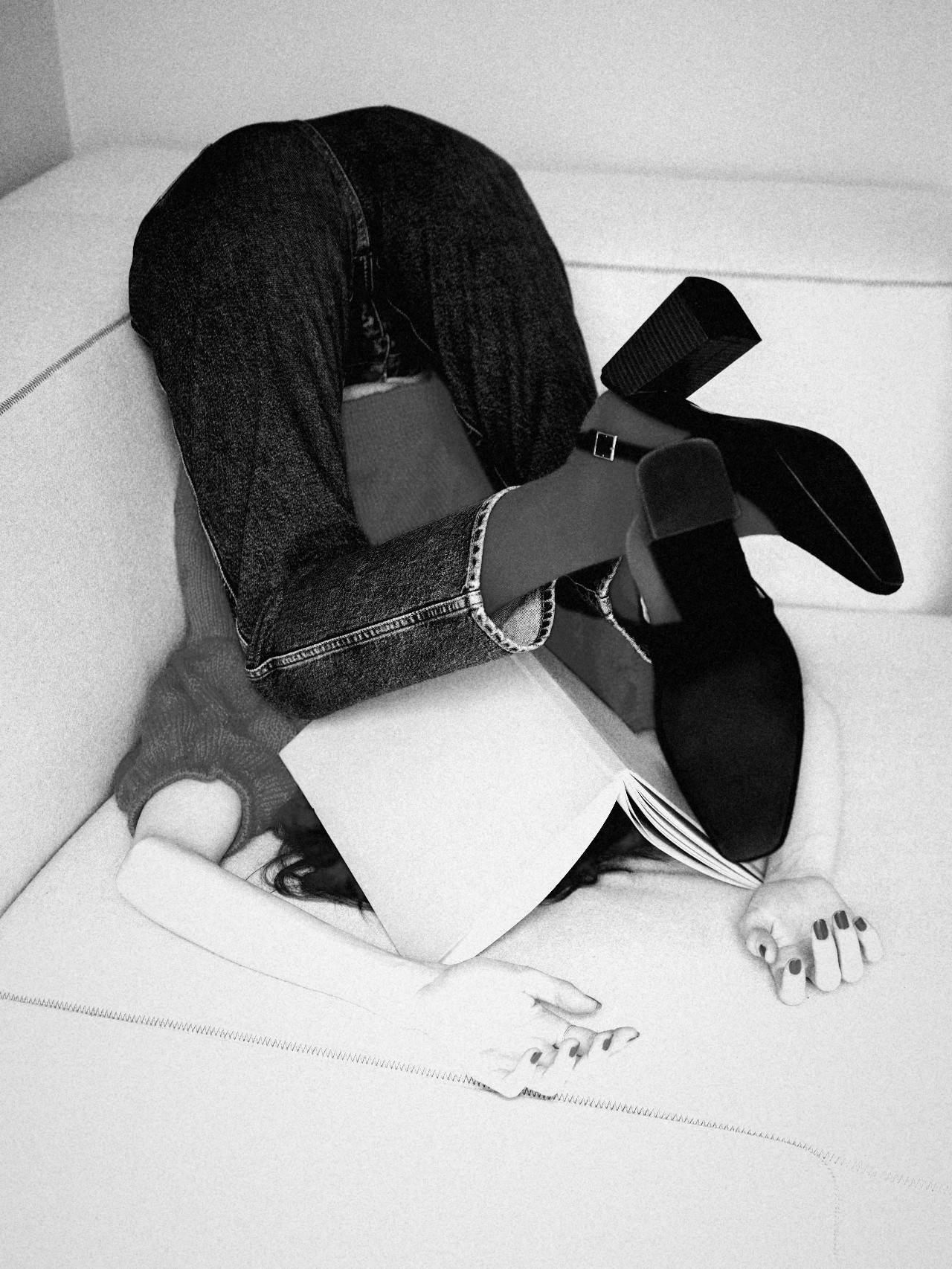A woman folded upside down on a white sofa, an open book over her face, black and white