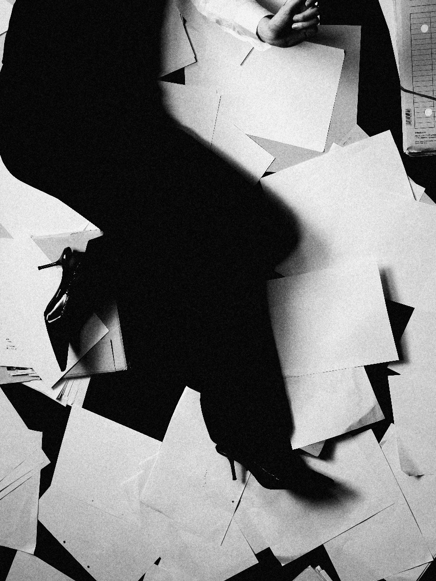 Seen from above: a woman in heels walking across a floor of loose papers, holding a phone cord, black and white
